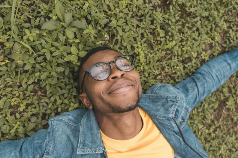 A person with glasses, wearing a yellow shirt and denim jacket, is lying on a patch of grass. They have their arms spread out and are smiling contentedly, looking upwards with a relaxed expression.