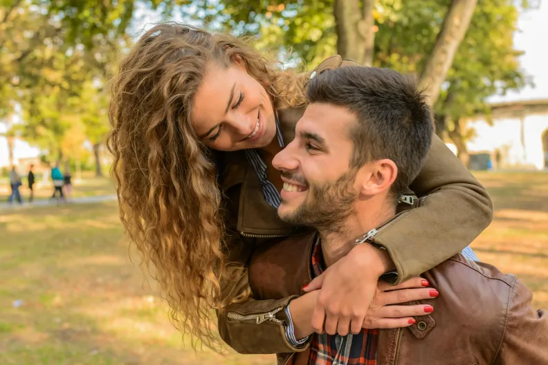 A woman with curly hair smiles as she hugs a bearded man from behind. They are outdoors in a park, surrounded by trees. The man, wearing a brown jacket, looks up at her with a joyful expression.