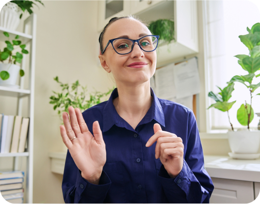 A person with short hair and blue glasses, wearing a blue shirt, smiles and holds one hand up as if waving. They are seated at a desk in a bright room with several green potted plants and a bookshelf in the background.