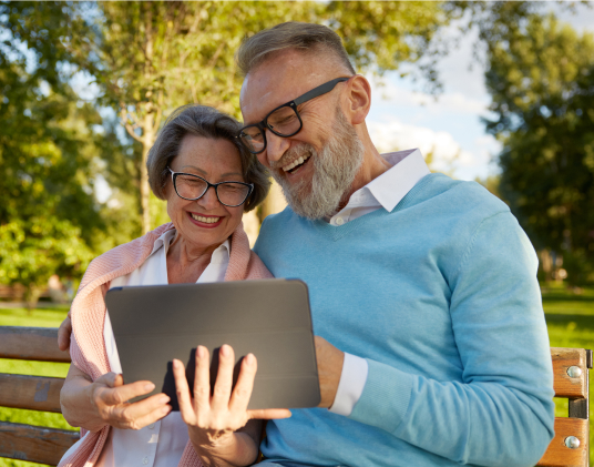 A senior couple sits on a bench in a park, smiling and closely looking at a tablet screen together. They both wear glasses and light clothing, enjoying a pleasant, sunny day outdoors with greenery in the background.